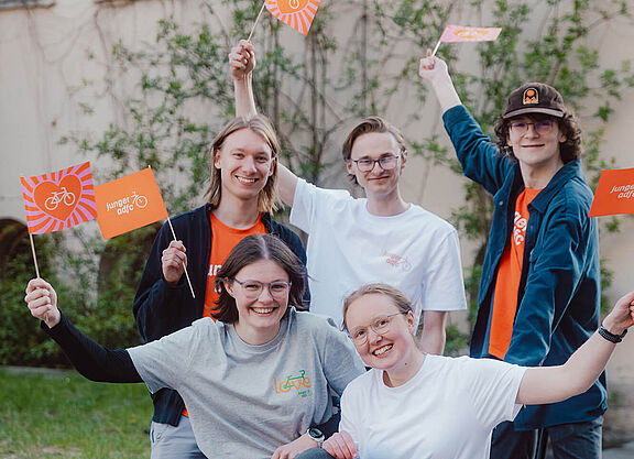 Bundesjugendvorstand Fünf junge Menschen posieren draußen auf und neben einem Lastenrad. Sie lachen und halten kleine Fahnen des Jungen ADFC in die Höhe.