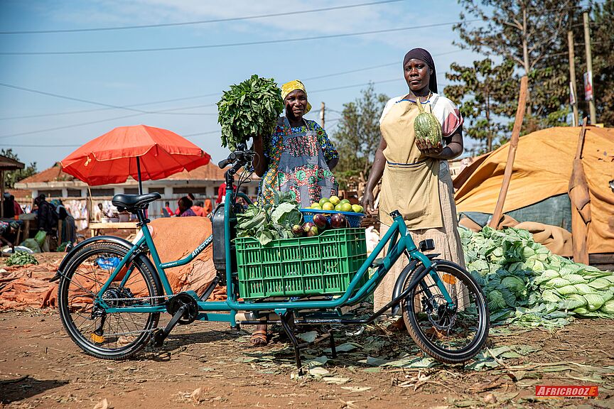 Markthändlerinnen mit AfricroozE-Lastenrad Zwei afrikanische Frauen stehen auf einem Markt im Freien hinter einem türkisgrünen Lastenfahrrad. Auf der tiefen Ladefläche zwischen Lenker und Vorderrad steht eine grüne Kiste, die voll mit Auberginen, Orangen und Blattgemüse beladen ist. Die Frauen halten weiteres Gemüse in den Händen und blicken freundlich in die Kamera.