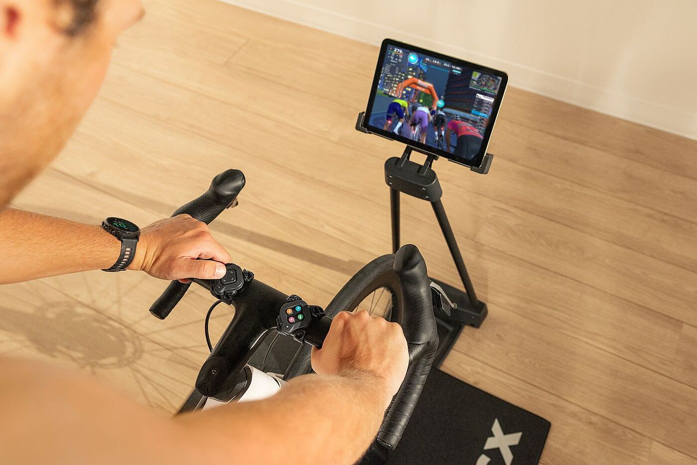 Hände liegen an einem Rennradlenker. Rechts steht ein Tablet auf einem Ständer, auf dem eine Gruppe Radfahrender zu sehen ist."
